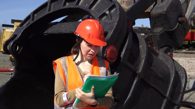 A young woman worker in an orange vest stands near a large ladle of a quarry excavator, looking over project.