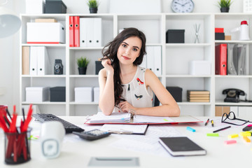 A beautiful young girl sits at the office table, holds her hair in her hand and looks straight.