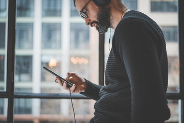 Closeup of handsome bearded man standing and using mobile phone for listening digital audio book. Horizontal.Panoramic windows on Blurred background