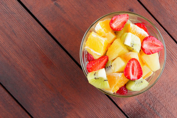 Fruit salad from pineapple, kiwi, orange, strawberry. Multicolored sliced fruit in a transparent dish. Vegetarian food on a wooden background. Copy space. Citrus fruits for Asian cuisine