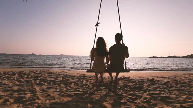 romantic couple in love sitting together on rope swing at sunset beach, silhouettes of young man and woman on holidays or honeymoon