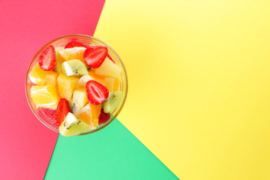 Fruit salad from pineapple, kiwi, orange, strawberry. Multicolored sliced fruit in a transparent dish. Vegetarian food on a multi-colored pop art background. Copy space.