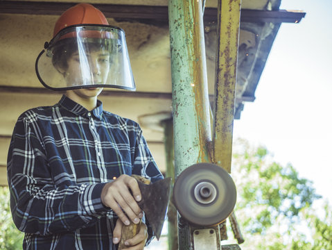 Young Teenage Man In Protactive Mask Working At The Grinder With Axe In The Hand