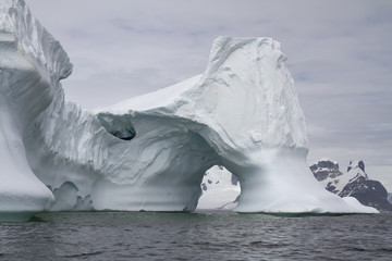 iceberg with an arch in Antarctic waters against the backdrop of the mountains of the Antarctic Peninsula on a cloudy day