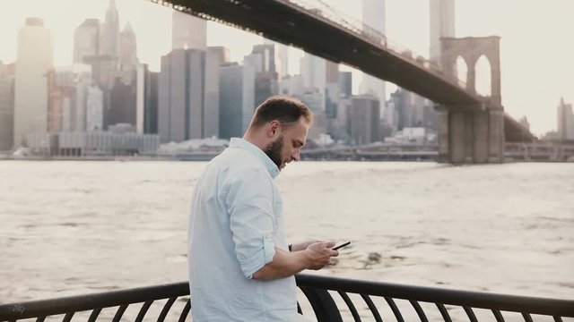 Happy European male tourist comes up to river embankment near Brooklyn Bridge, New York, takes photo and smiles 4K.