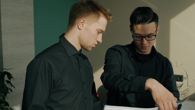 Young male colleagues are talking standing in modern office, handsome guys in black shirts discussing new project, working, smiling at company. Concept: collaborating, business communication