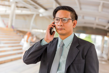 Young Asia handsome businessman with his smartphone standing on walkway of modern city.