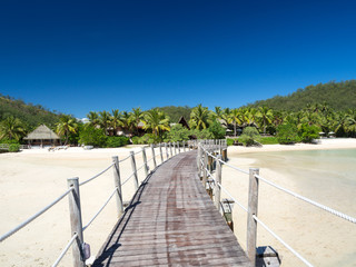 Obraz premium Beautiful Luxury Boardwalk Walkway Over Beach to Palm Tree Island with Bure Bungalows on The South Pacific Tropical Island of Fiji