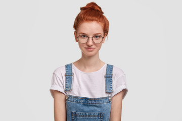 Pretty female wonk student, has healthy skin, red hair, wears denim overalls, waits for friend to go together in library, smiles pleasantly, likes studying and reading, isolated over studio wall