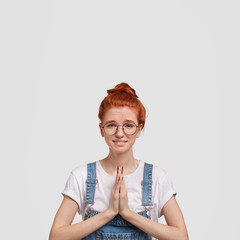 Please, forgive me! Adorable freckled female has sorrowful expression, pleads for forgiveness, keeps palms together, feels guilty, did something wrong, poses indoor against blank white wall.