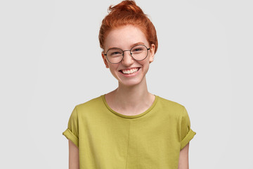 Photo of tender delighted female student with freckled skin, foxy hair and broad smile, wears spectacles and casual green t shirt, has fun with groupmates during break at university or college