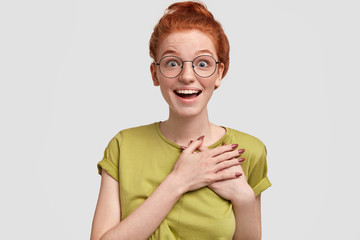 Excited ginger female keeps hands on heart, feels surprised as recieves unexpcted news, wears spectacles, poses against white background. Ginger freckled girl expresses excitement with present