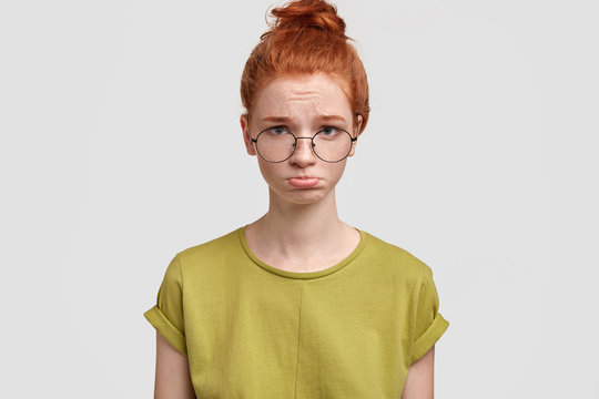 Portrait of discontent female frowns face in discontent, looks desperately at camera, going to cry, has freckled skin, poses against white studio wall. Redhead woman cries and being offended