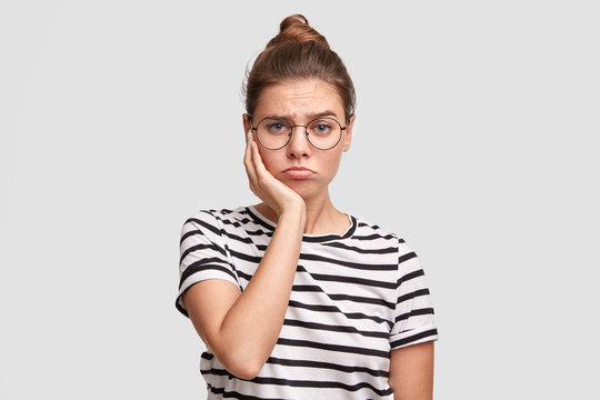 Photo of serious unhappy European female keeps hand on cheek, has sullen expression, wears spectacles and casual striped t shirt, has desperate expression, recieves bad news, keeps hand on cheek