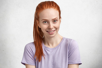 Close up shot of pleasant looking ginger young female smiles gladfully, wears purprle t shirt, looks directly at camera, poses against white concrete wall, listens friend`s interesting story