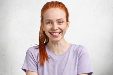 People, beauty and skin care concept. Pretty freckled female with ginger hair combed in pony tail, dressed in casual clothing, poses against white background. Red haired woman rejoices new purchase