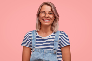 Obraz premium Glad smiling pretty female freelancer, has broad smile, demonstrates teeth with braces, wears casual t shirt with overalls, rejoices recieving reward, stands against pink background. Emotions concept