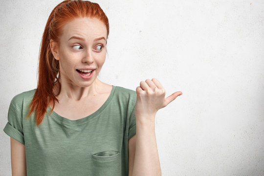 Portrait of attractive female with freckled female has foxy hair tied in pony tail, dressed in casual t shirt, points aside at blank copy space, has excited expression. People, advertisement and fear