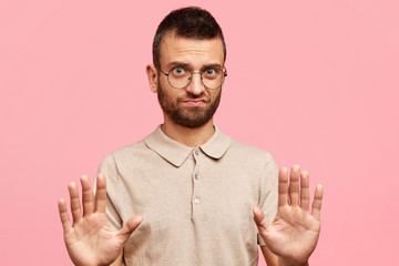 Serious unshaven male student shows refusal gesture, rejects going to blackboard and give answer on question, frowns face, wears glasses, isolated on pink background. Don`t bother me, please!