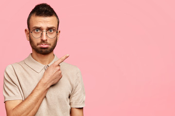 Worried displeased young male with trendy haircut, bristle, wears casual t shirt and round spectacles, indicates with fore finger at blank space, shows something scary, isolated on pink wall
