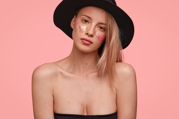 Pleasant looking young female with naked shoulders, has glitters on cheek, shows her artistic make up, prepares for carnival, wears black elegant hat, poses against pink background. Fashion concept