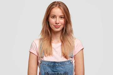 Obraz premium Attractive teenager with confident look, has long straight hair, dressed casually, smiles gently, looks directly at camera, reads necessary information in front, stands against white concrete wall