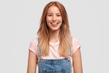 Obraz premium Positive glad delighted female student has broad smile and happy look, rejoices coming weekend, has plans to go on picnic with friends, dressed in denim overalls. People and positive emotions