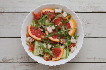 Grapefruit salad with chicken breast, fresh arugula leaves, red cherry tomatoes, cucumber in white salad bowl on wooden table. Balanced meal