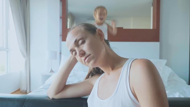 Upset mother with hyperactive child jumping on bed on the background