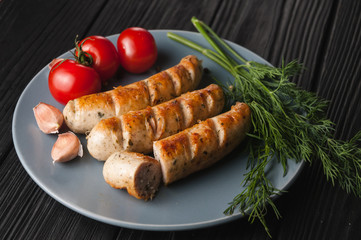 Three grilled sausages on a gray plate on a wooden black background with cherry tomatoes, herbs, garlic.