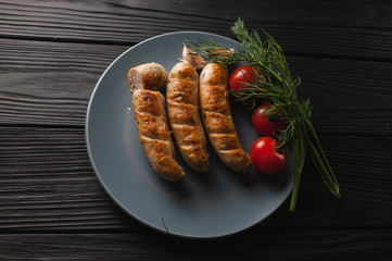 Three grilled sausages on a gray plate on a wooden black background with cherry tomatoes, herbs, garlic.