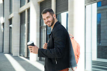 cheerful attractive man with a beard holds a paper Cup with coffee on his outstretched hand. The positive expression of the sun and the environment of the streets create an atmosphere of good morning.