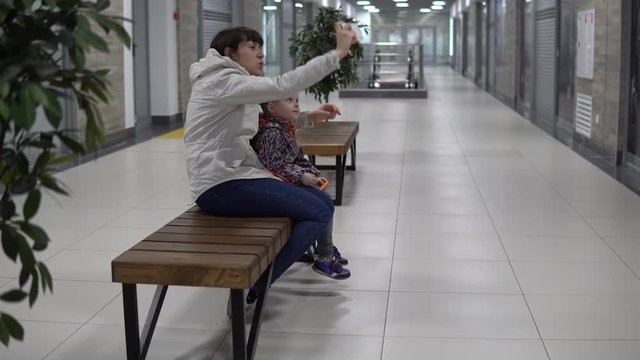 Mom with daughter are doing selfie or make a video call using the smartphone, the family shares their good mood, sitting on a bench in the mall.