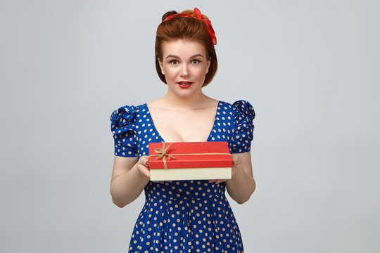 Picture of attractive glamourous young Caucasian woman wearing bright make up and elegant blue dotted dress, holding fancy red box, looking at camera, handing it over to you as birthday present