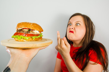 Young fat woman shows middle finger aside the burger on gray background