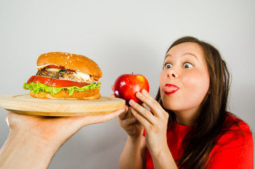 Young fat woman holds red apple, looks at the burger and shows tongue on a gray background.
