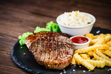Grilled steak with french fries and vegetables served on black stone on wooden table