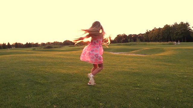 Cute schoolchild spinning on green meadow. Slow motion joyful little girl having fun on green lawn, sunset sky. Active leisure on fresh air.