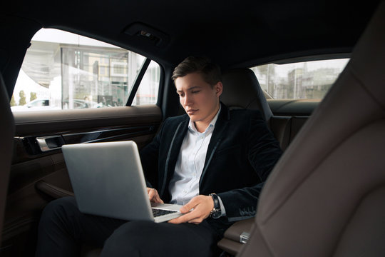 It is time to work! Handsome good-looking young busy man seriously doing his work on the laptop on the rear seat in the car.