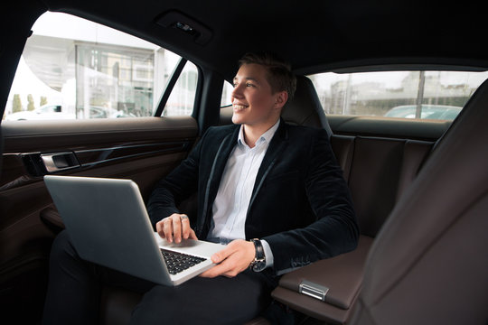 Cheerful work! Smart clever young smiling newcomer businessman using internet, while working with laptop in the automobile.