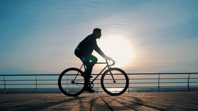 Close up shot of man's silhouette riding road bike on the wooden beach during sunset or sunrise. Boy with his bicycle. Traveler. Healthy lifestyle concept. Slow motion.