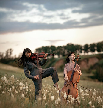 Female Musical Duet With Violin And Cello Plays On Flowering Meadow Against Backdrop Of Picturesque Landscape.