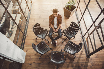 Top view full length portrait of unhappy bearded male sitting opposite empty chairs indoor. Loneliness concept © Yakobchuk Olena