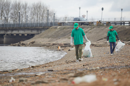 Two young men in green uniform walking along waterside and seeking for litter to pick it up