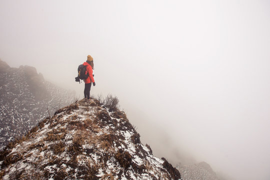 Young traveller woman wearing backpack, red jacket and hat standing in fog mountain trekking above clouds and valley. No way, lost in mountains