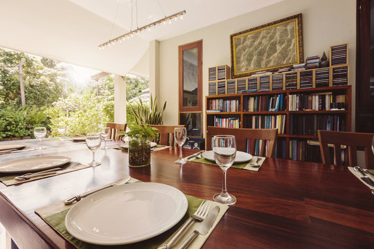 Luxury Tropical dining room with bookshelf, dvd collection, open outdoor space. Plates and vine glasses on wooden table, beautiful summer villa design