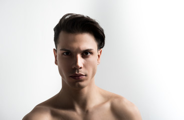 Portrait of youthful shirtless man with perfect and pure skin. He is standing against light background and looking at camera thoughtfully. Copy space in the right side. Skincare concept