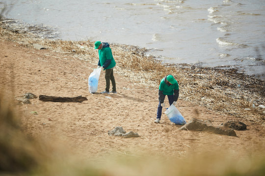 Two youn men in green uniform cleaning river bank from litter and putting it into sacks