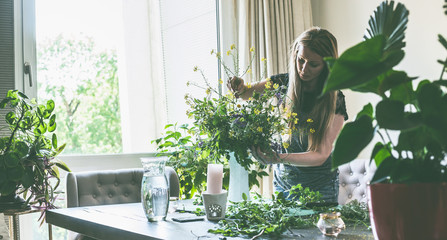 Pretty woman with long hair arranging wild flowers bunch in vase on table in living room at window...