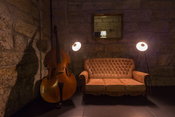 leather sofa, cello music instrument, lamps in dark interior. Stone wall on background, brutal...